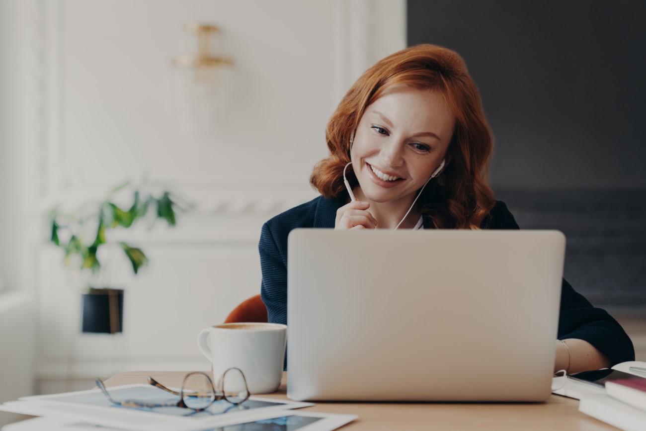 Positive ginger businesswoman works remotely from home during quarantine, has video conference with colleagues, makes online call, uses wireless internet and modern laptop computer with earphones