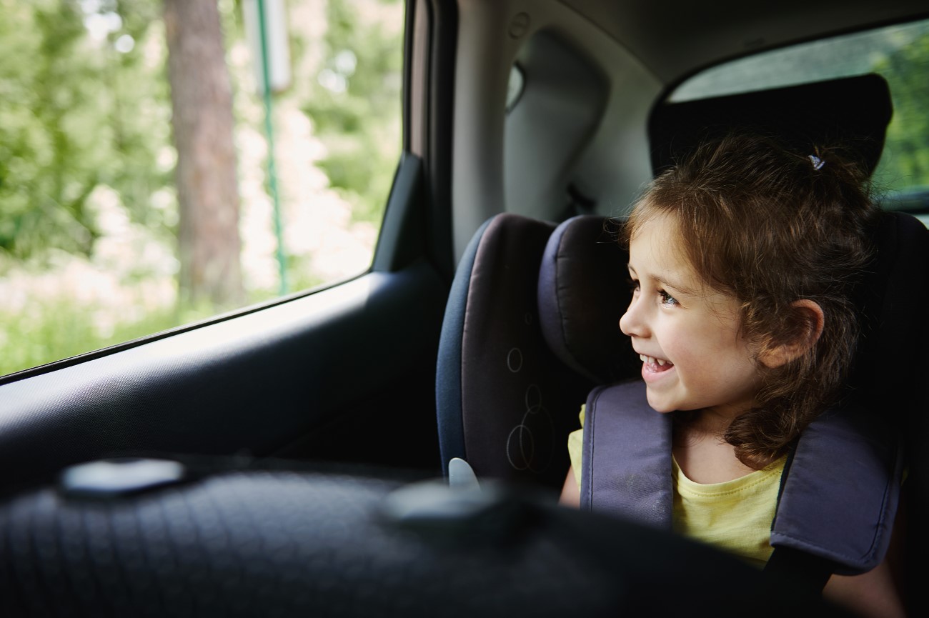 Adorable baby girl enjoys the travel by car, looks out of an opened window while sitting in a safety booster car seat